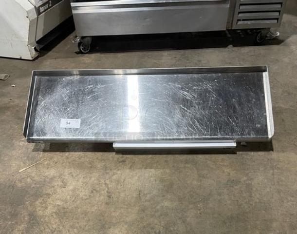 Commercial stainless steel wall-mount shelf with ticket holder, rectangular design. Slight surface scratches, durable and functional.