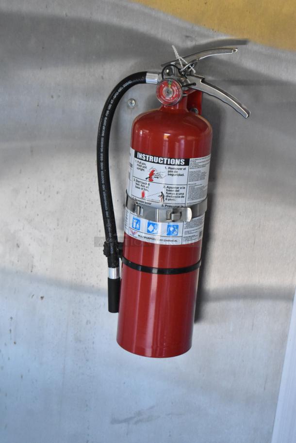 Red fire extinguisher mounted on a metal wall, featuring safety instruction label and black hose, part of a mobile kitchen.