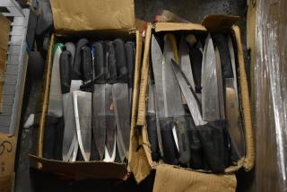 85 sharpened stainless steel knives with black handles, including a chef's knife, in worn cardboard boxes.
