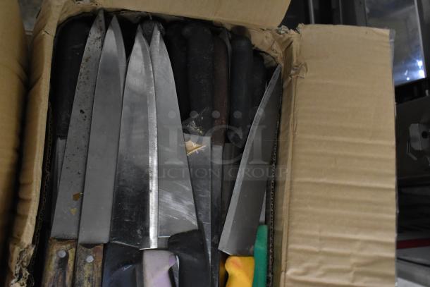 Box of assorted sharpened stainless steel knives, including chef knives, showing varied handles and blade conditions.