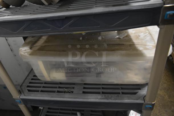 Two bins with assorted ice cream machine parts, including augers and a front panel, stored on a gray shelving unit.