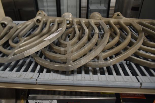 Nine Hobart mixer paddle attachments, metal, slightly used, arrayed on a grate shelf. Visible tag with auction info.