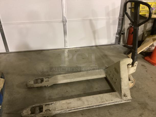 Tan pallet jack with black handle, dual forks, and rolling wheels. Used condition, ideal for warehouse lifting tasks.