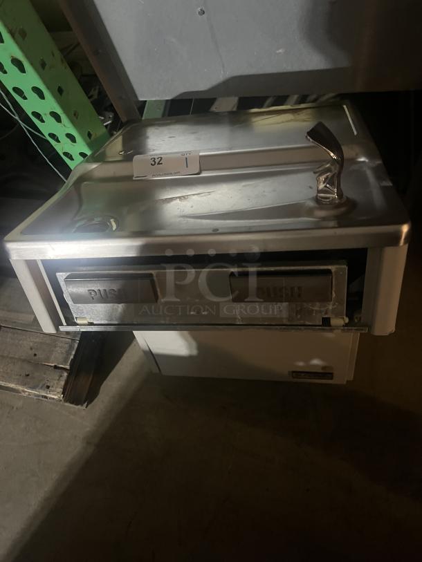 Stainless steel water fountain with dual push bars and bubbler, mounted on a light-colored base. Marked with auction label "32".