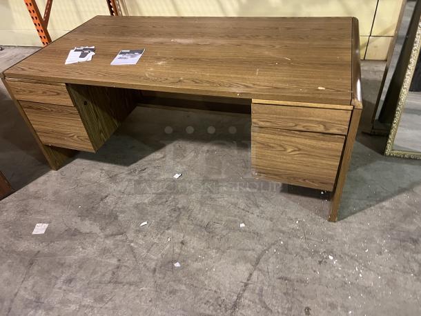 Wooden desk with a laminate finish, featuring two side drawers. Minor surface wear visible. Includes paper sheets on top.