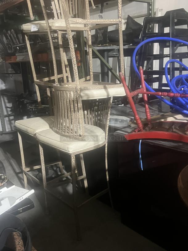 Two wrought iron style barstools with cushioned seats, showing ornate design. Some visible wear, stacked in storage.