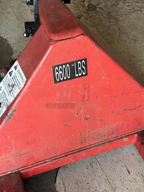 Red pallet jack with a load capacity of 6600 lbs. Visible scratches indicating moderate wear. Warning label attached.
