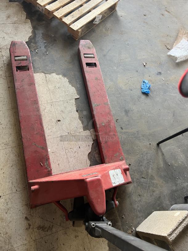 Red pallet jack with visible wear, showing surface scratches. Includes as-is label and handle, suitable for heavy lifting.