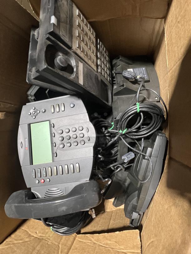 Polycom hard-wired office phones in a cardboard box, featuring numeric keypads, LCD screens, and coiled cords. Used condition.