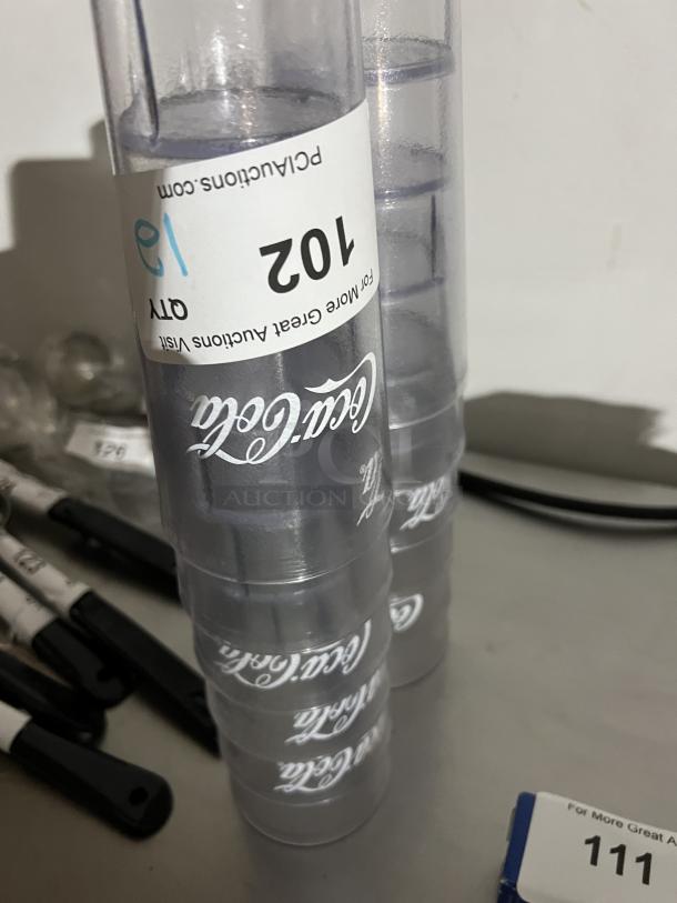 Coca Cola clear plastic cups, set of 12, stacked with visible logo, in good condition. Label reads "Lot 102, Qty 12".