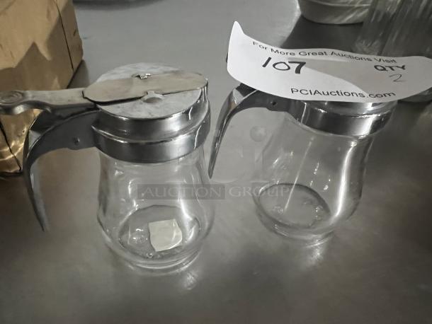 Two clear glass syrup dispensers with metal lids and handles, in good condition. Auction tag marked "107, Qty 2."