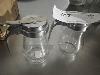 Two clear glass syrup dispensers with metal lids and handles, in good condition. Auction tag marked "107, Qty 2."