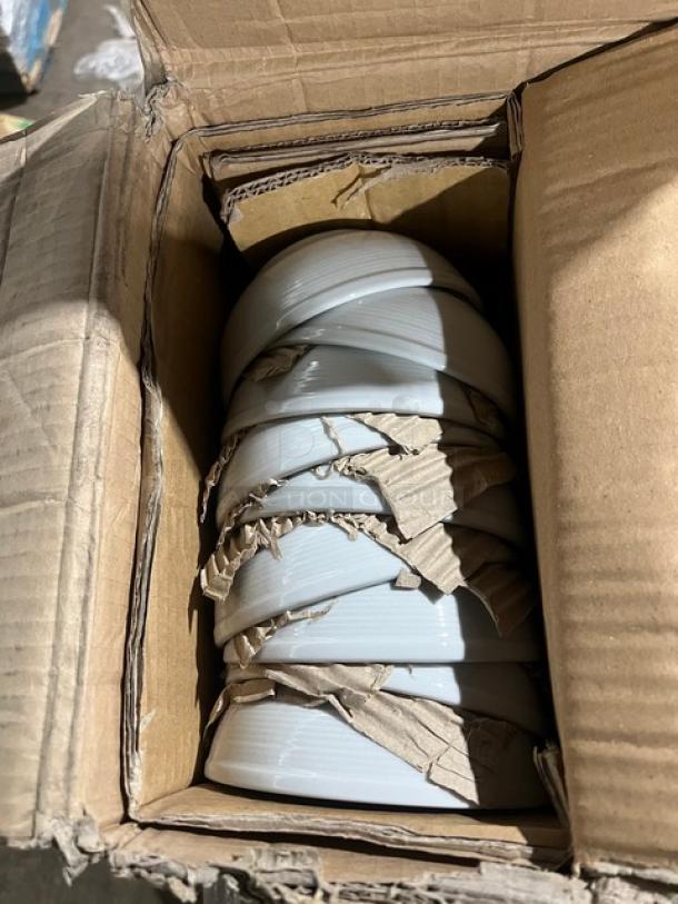 Box of stacked white porcelain plates, partially wrapped in cardboard for protection, visibly new and intact.