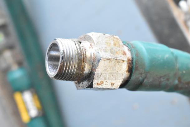 Close-up of a metal gas connector with threading, showing signs of wear and rust, attached to a green hose.