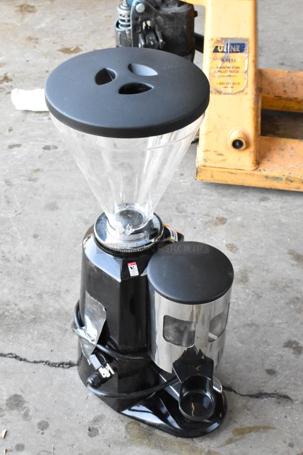 Estella espresso machine shown on ground, featuring black and silver design, with visible buttons and a black power cord.