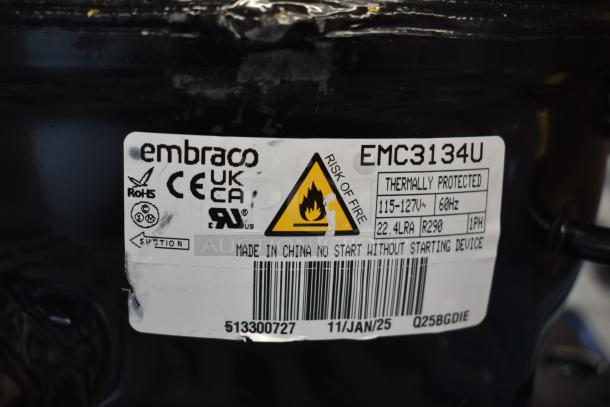 Brand new Embraco EMC3134U compressor, 115V, R-290, thermally protected. Tags show certifications, model, and safety warnings.