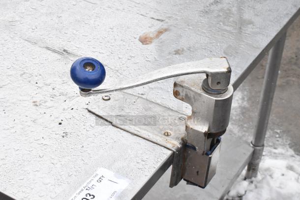 Metal table with attached commercial can opener, blue handle, and under shelf. Visible wear and rust on the opener.