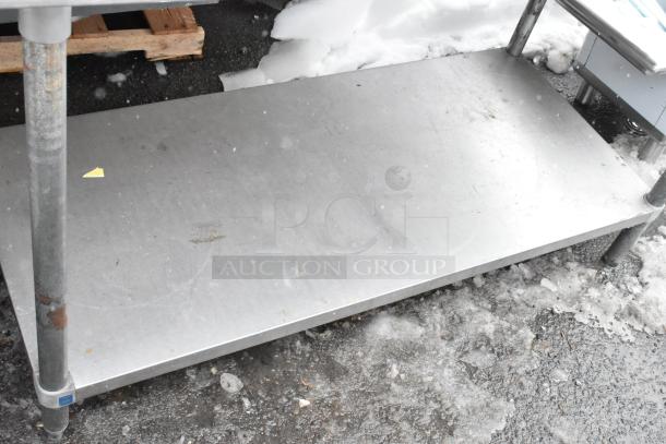 Stainless steel table with under shelf, industrial style, sturdy metal legs, some surface wear visible, outdoor setting.