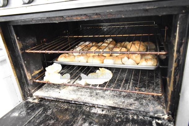 Bakers Pride commercial gas range with 6 burners, flat top griddle, cheese melter, 2 ovens, and backsplash. Ovens show signs of use.