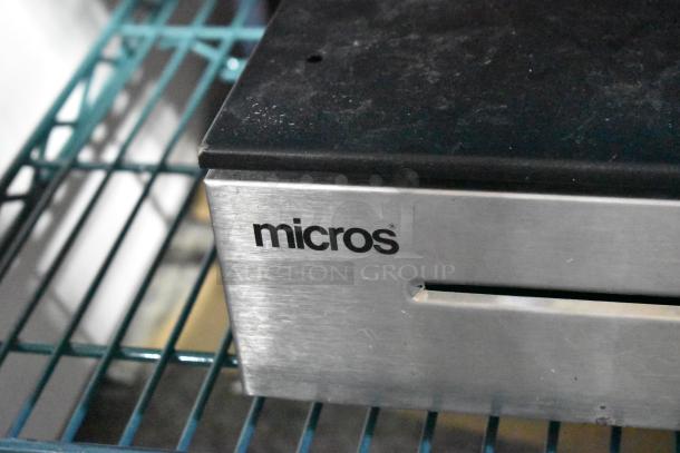 Micros cash drawer with stainless steel front, visible slot, and "micros" branding. Shows signs of use, resting on a blue wire rack.