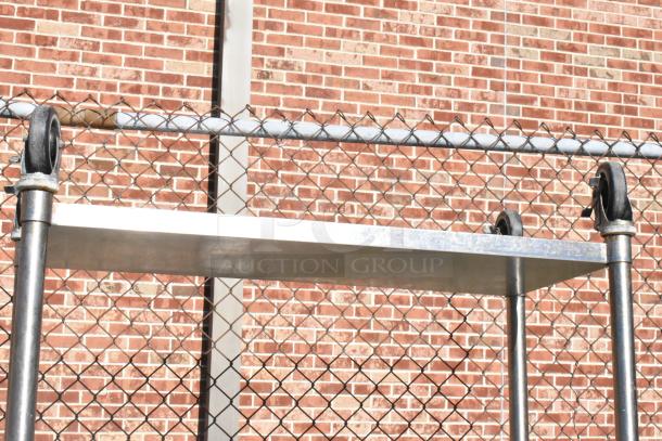 Stainless steel commercial table with under shelf and casters, near chain-link fence; good condition, industrial use.