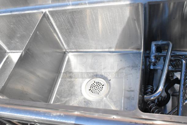Commercial 3 bay stainless steel sink with dual drain boards, featuring visible drain and plumbing components, in used condition.