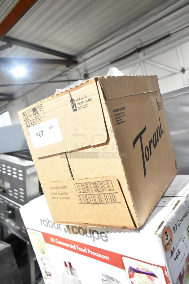 Cardboard box labeled "Torani" for 12 x 25.4 fl oz glass bottles, marked "scratch and dent," atop a Robot Coupe food processor box.