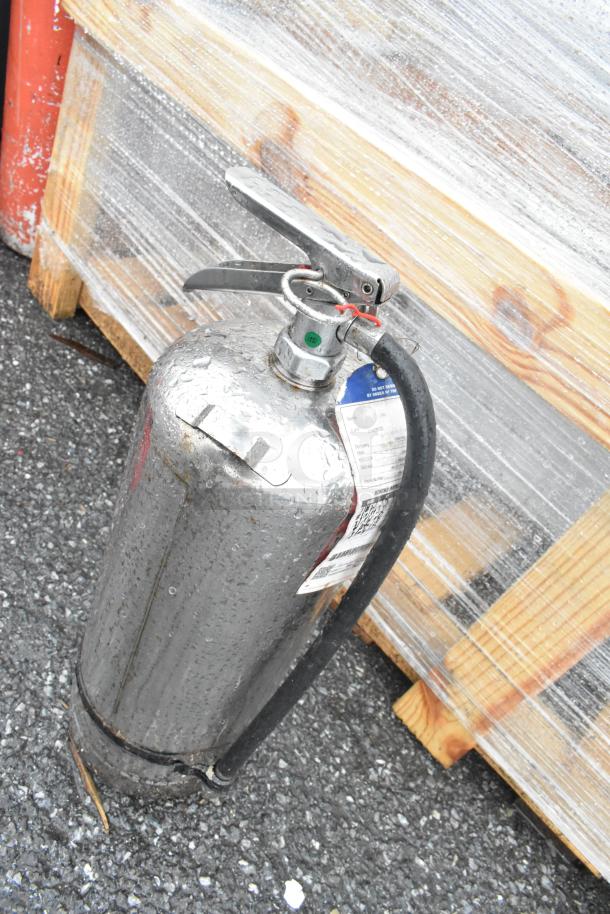 Two stainless steel fire extinguishers with hoses, outdoor condition, attached inspection tags. Buyer pickup required.