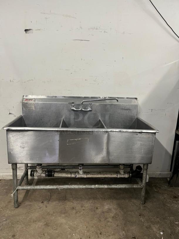 Stainless steel 3 compartment sink with backsplash, visible wear, sturdy frame, and integrated plumbing. Ideal for commercial kitchens.
