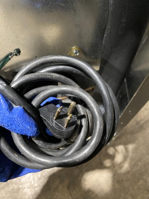 Close-up of a power cord plug for a Delfield Model 406CADD1 stainless steel undercounter refrigerator, held by a gloved hand.