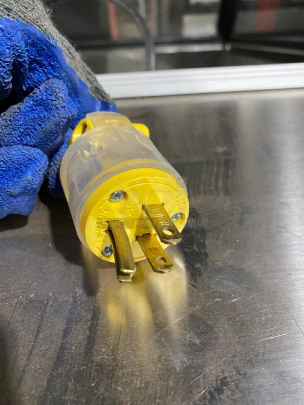 Yellow electrical plug with three prongs, set on a metallic surface. Blue glove partially visible.