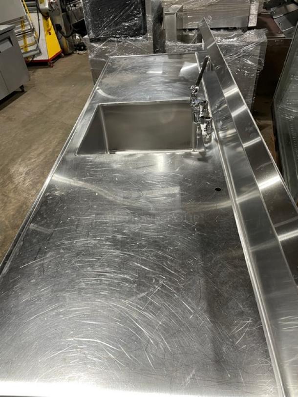 Custom commercial stainless steel sink worktop table with backsplash and swivel spout faucet. Features dual-sided undershelf and legs. Slight wear visible.