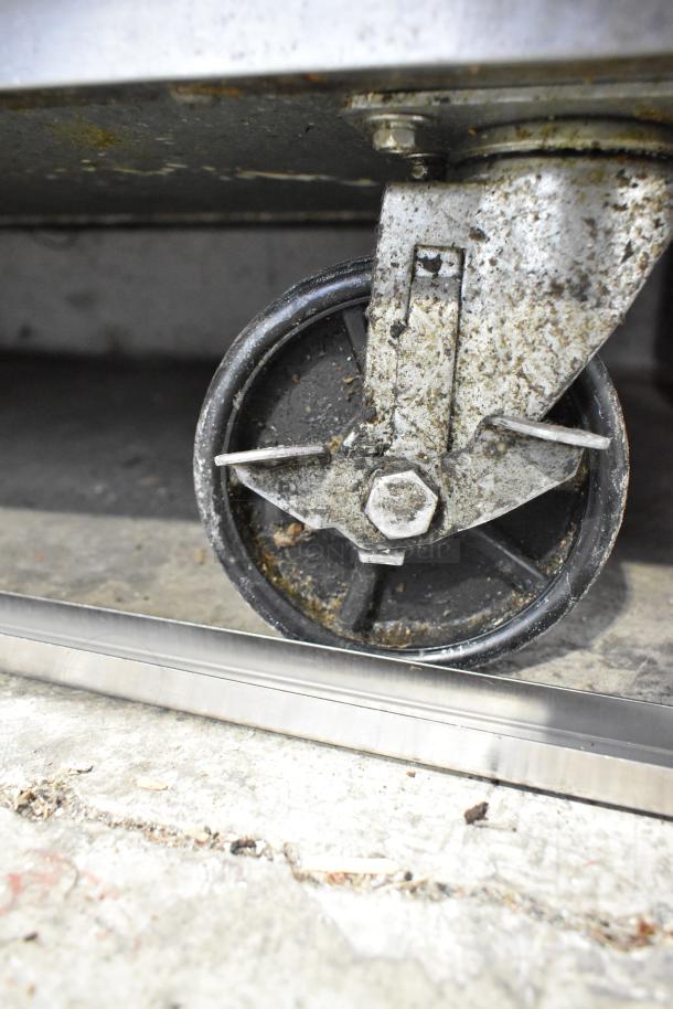 Dukers prep table caster wheel detail, showing wear and sturdy construction with visible grime, on a metallic finish.