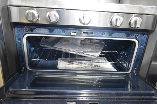 Gas-powered 4-burner range with two ovens, missing grates. Knob controls, open oven showing interior with manuals.