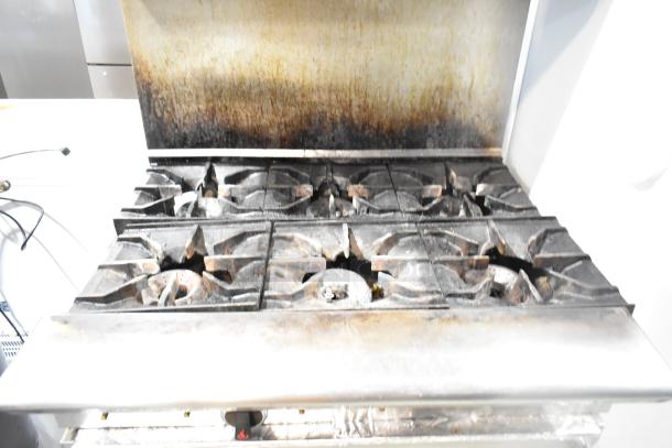 American Range commercial gas-powered 6 burner range with oven, over shelf, and backsplash. Signs of use on burners.