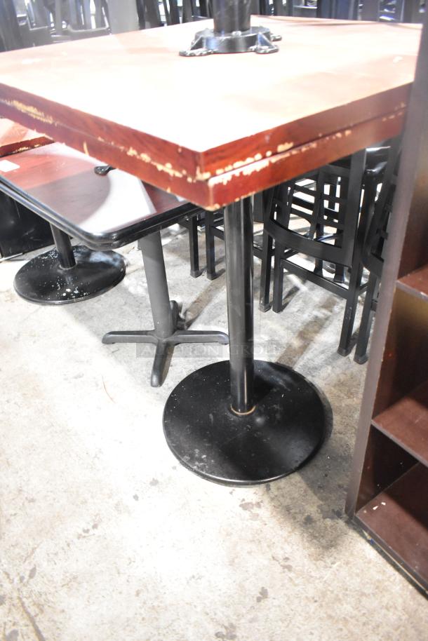 Two bar height tables with wood pattern tabletops and black metal bases, showing signs of wear.