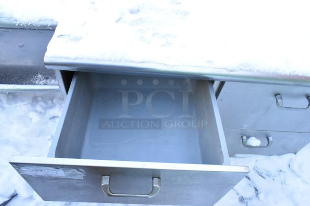 Stainless steel work table with four drawers and backsplash, featuring sturdy handles. Snow-covered, showing one open drawer.