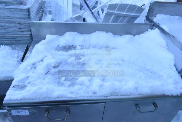 Stainless steel work table with four drawers and backsplash, partially covered in snow. Suitable for heavy-duty use.