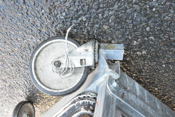 Metal pan transport rack with sturdy commercial casters, showing signs of wear, on asphalt surface.
