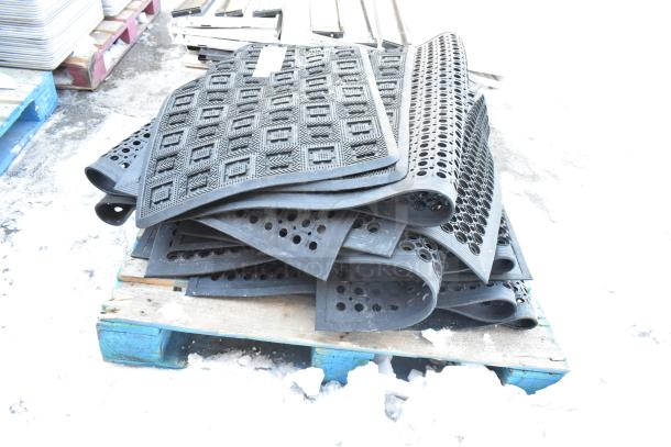 14 assorted black rubber floor mats with different patterns and perforations, stacked on a wooden pallet.
