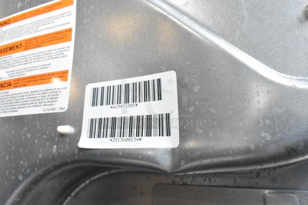 Barcode and warning labels on Frigidaire FLCE7522AW stacked washer and dryer, gray surface, minor scratches visible.