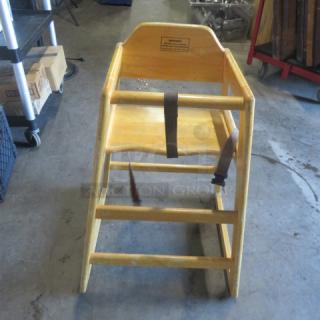 Wooden high chair with safety straps, natural finish, visible wear. Features secure seating with mounted safety labels.