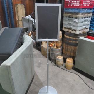 Metal dual-sided sign on stand, silver frame, adjustable height, located in a warehouse setting. Good condition.