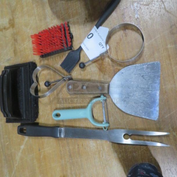 Lot of miscellaneous kitchen and barbecue tools including spatula, peeler, pastry brush, and grilling fork on wooden surface.