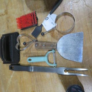 Lot of miscellaneous kitchen and barbecue tools including spatula, peeler, pastry brush, and grilling fork on wooden surface.