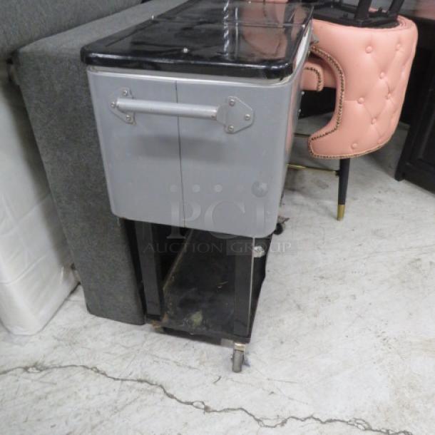 Portable beverage/ice cooler with flip lid, undershelf, and bottle opener on casters. Gray finish, sturdy condition.
