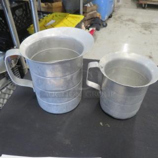 Assorted aluminum measuring pitchers, two sizes, with sturdy handles, in good condition, no labels or markings visible.