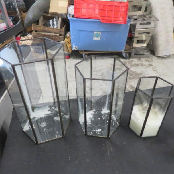 Set of 3 hexagonal glass candle holders of varying heights, black metal frames, some wax residue visible inside.