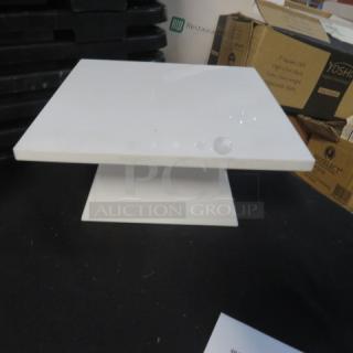 Square white cake display stand, 9.5x9.5x4.5 inches, in good condition. Suitable for showcasing cakes or food items.