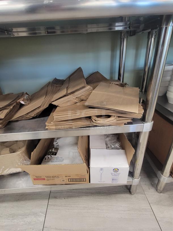 KCS WS-2460 Stainless Steel Work Table with bulk paper bags, drink carriers, and plastic cutlery displayed on shelves.